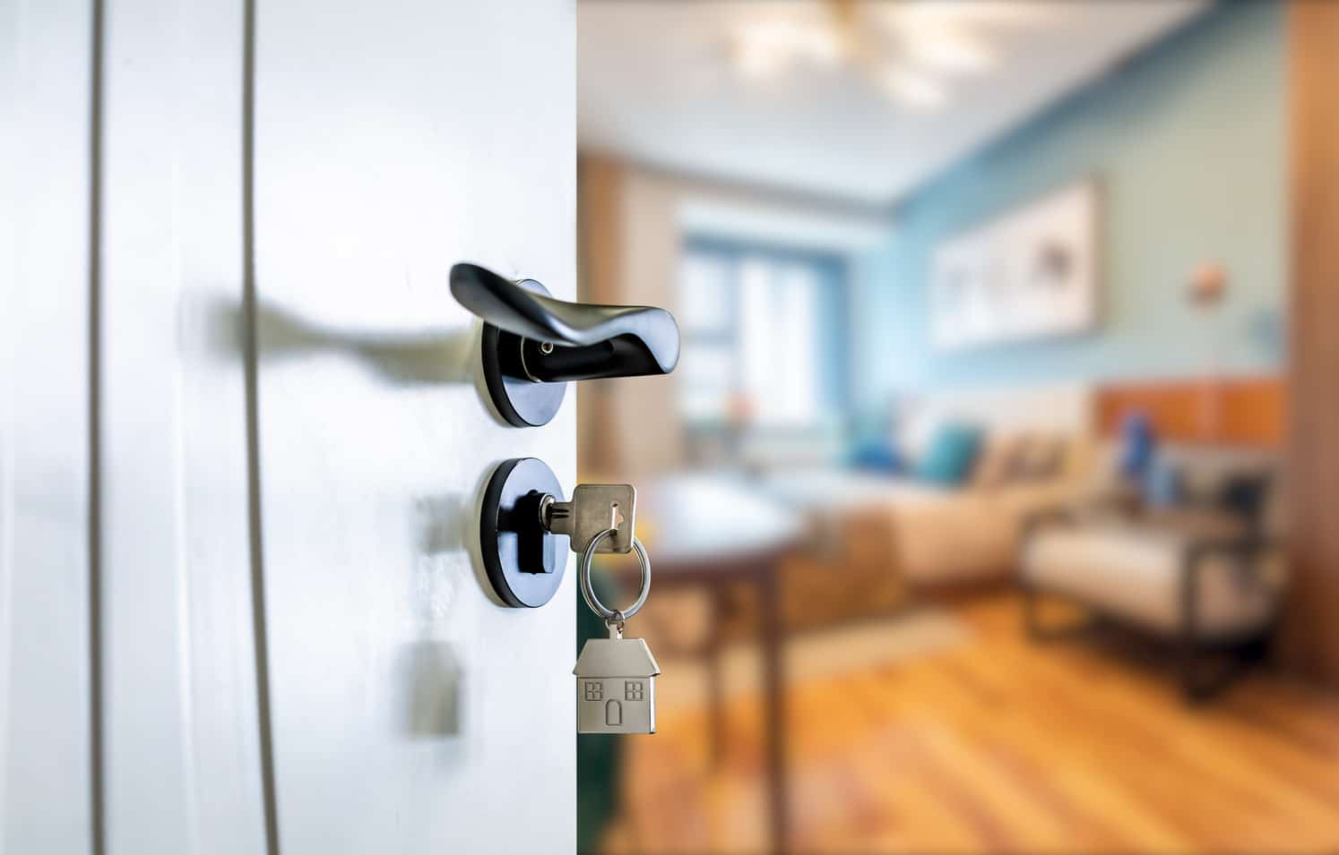 Close-up of an open door with a key in the lock, featuring a house-shaped keychain. A blurred living room is visible in the background, suggesting a perfect entry for estate agent websites to showcase property interiors.