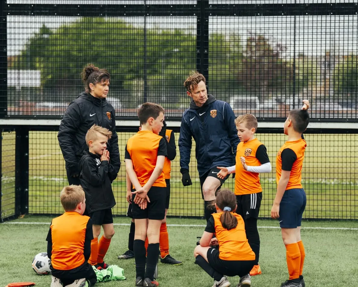 Two coaches talk to a group of young Wilvale Rangers soccer players in orange uniforms on an outdoor field during practice.