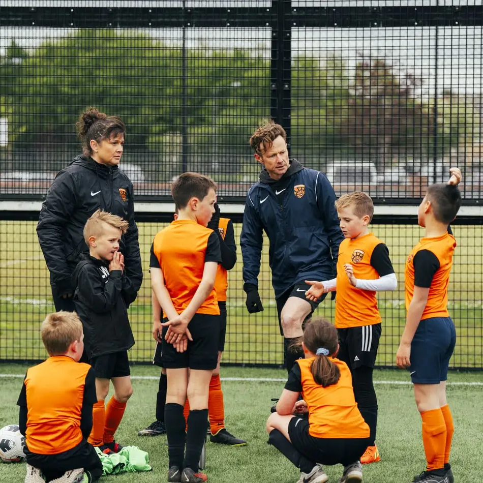 Two coaches talk to a group of young Wilvale Rangers soccer players in orange uniforms on an outdoor field during practice.