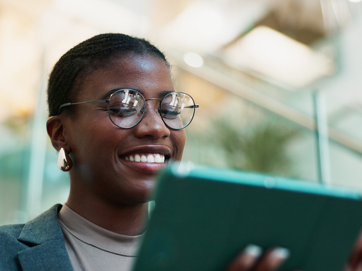 A person in glasses and a blazer smiles while using a tablet, standing in a bright indoor space with glass railings, likely planning the next steps for their website designing project.