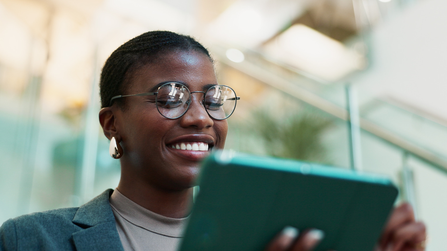 A person in glasses and a blazer smiles while using a tablet, standing in a bright indoor space with glass railings, likely planning the next steps for their website designing project.