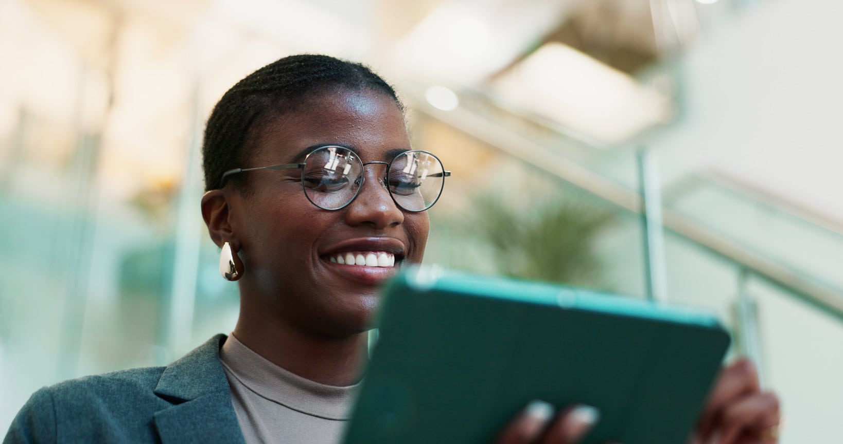 A person in glasses and a blazer smiles while using a tablet, standing in a bright indoor space with glass railings, likely planning the next steps for their website designing project.