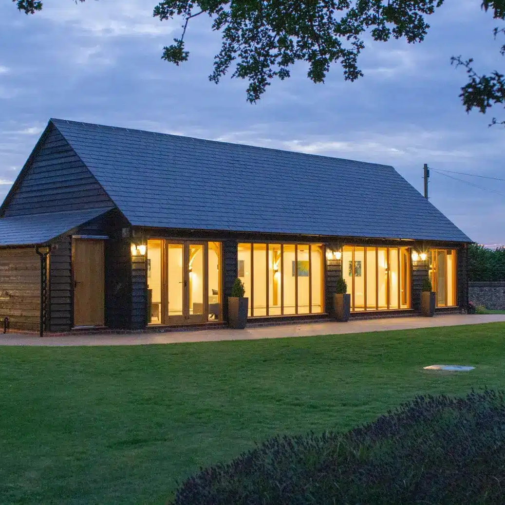 A modern barn-style cartlodge with large glass doors and exterior lights, surrounded by a well-kept lawn at dusk.