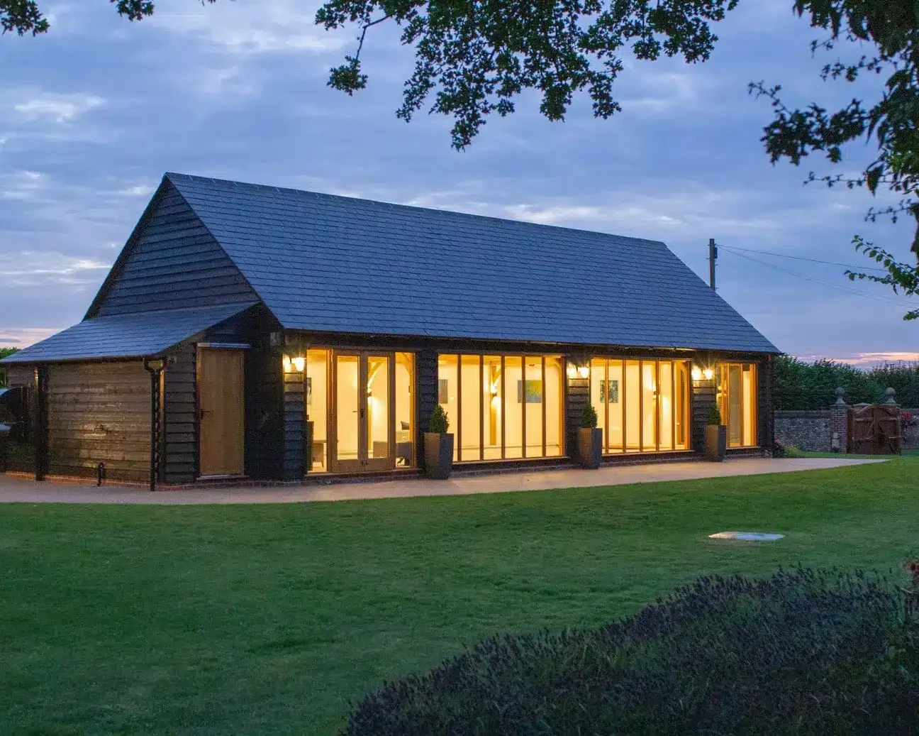 A modern barn-style cartlodge with large glass doors and exterior lights, surrounded by a well-kept lawn at dusk.