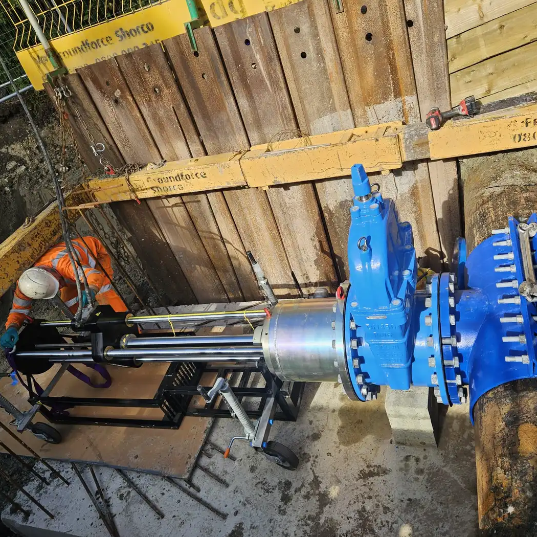 A worker operates machinery to install or maintain a large blue water valve on an underground pipeline at a construction site.
