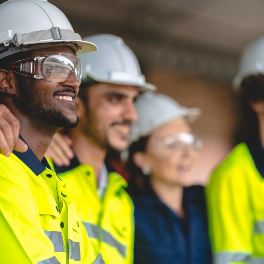 Four workers wearing safety helmets, high-visibility jackets, and safety glasses stand side by side, smiling and looking in the same direction.
