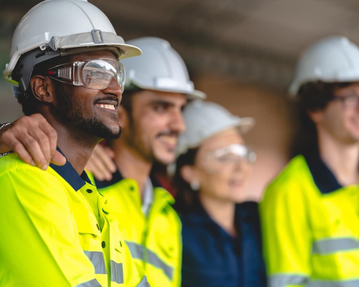 Four workers wearing safety helmets, high-visibility jackets, and safety glasses stand side by side, smiling and looking in the same direction.