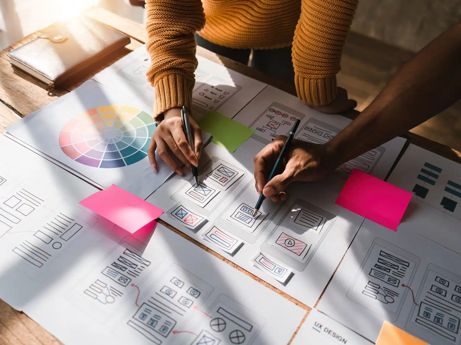 Two people work on website wireframe sketches at a wooden table covered with design papers, a color wheel, and sticky notes, exploring web design trends that could shape the future of web design.