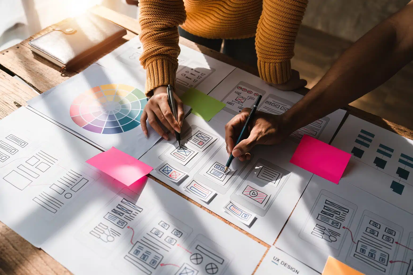 Two people work on website wireframe sketches at a wooden table covered with design papers, a color wheel, and sticky notes, exploring web design trends that could shape the future of web design.
