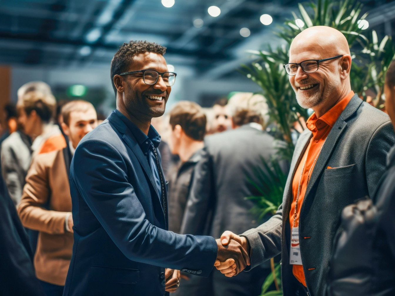 Two men in business attire smile and shake hands at the Chelmsford Business networking event, with others conversing in the background.