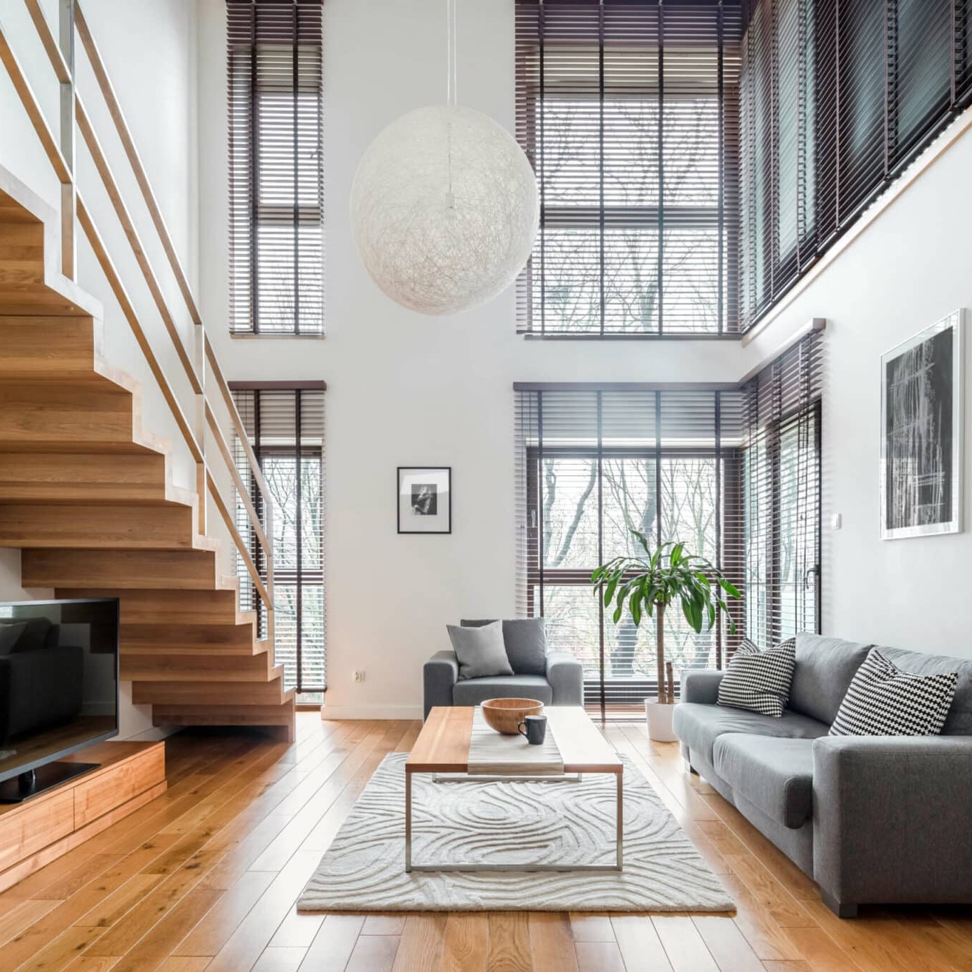 Modern living room with large windows, wooden staircase, gray sofa, coffee table, TV, and decorative plants—Balch Estate Agents present this bright space filled with natural light and minimalist decor.