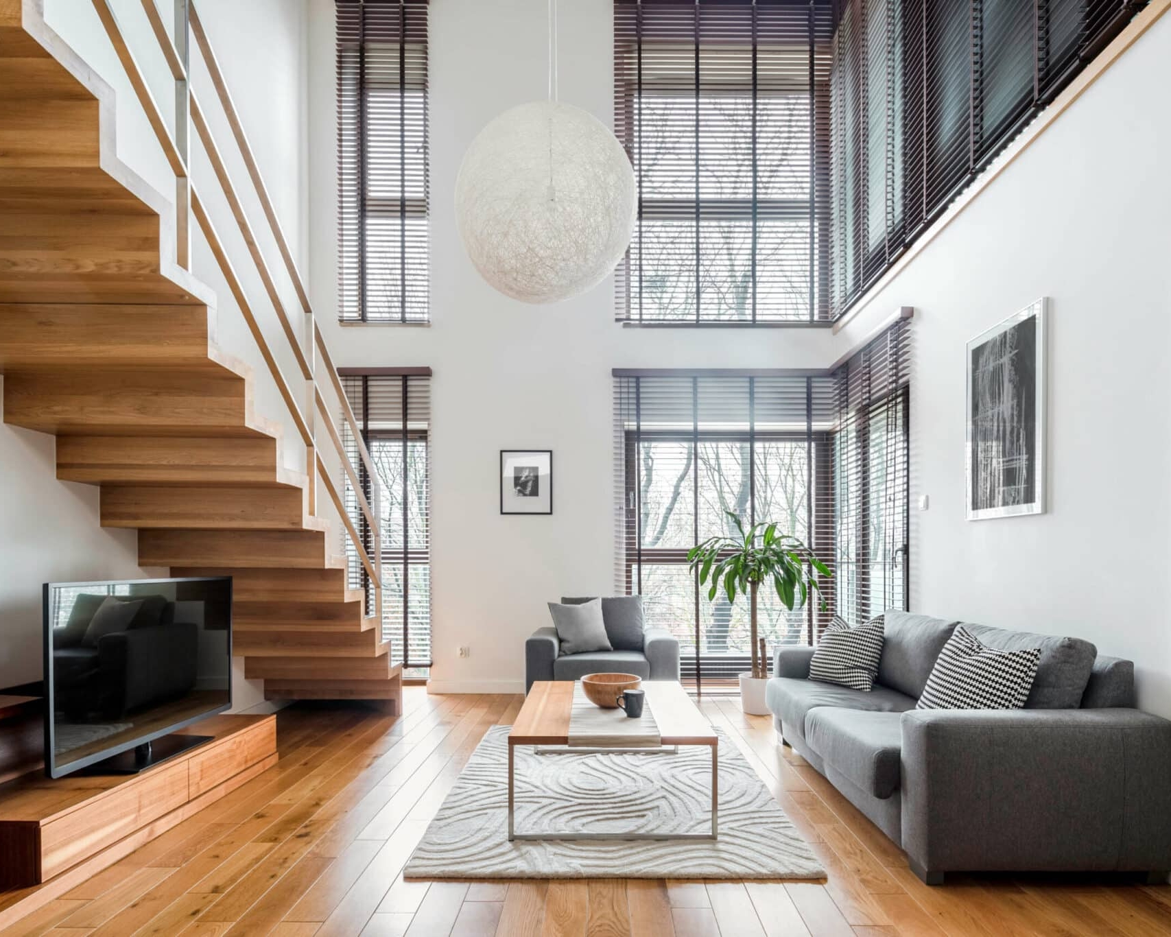Modern living room with large windows, wooden staircase, gray sofa, coffee table, TV, and decorative plants—Balch Estate Agents present this bright space filled with natural light and minimalist decor.