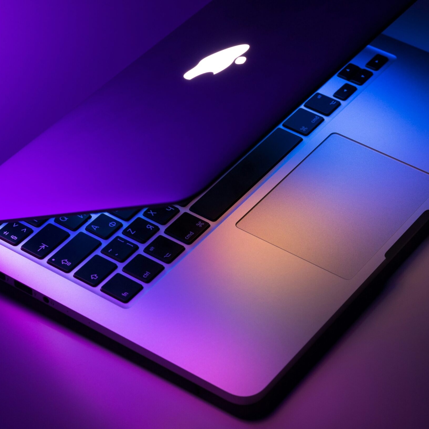 A partially open MacBook laptop with illuminated keyboard and trackpad, featuring purple and orange lighting reflections—showcasing the expert touch of Hardy Repairs.