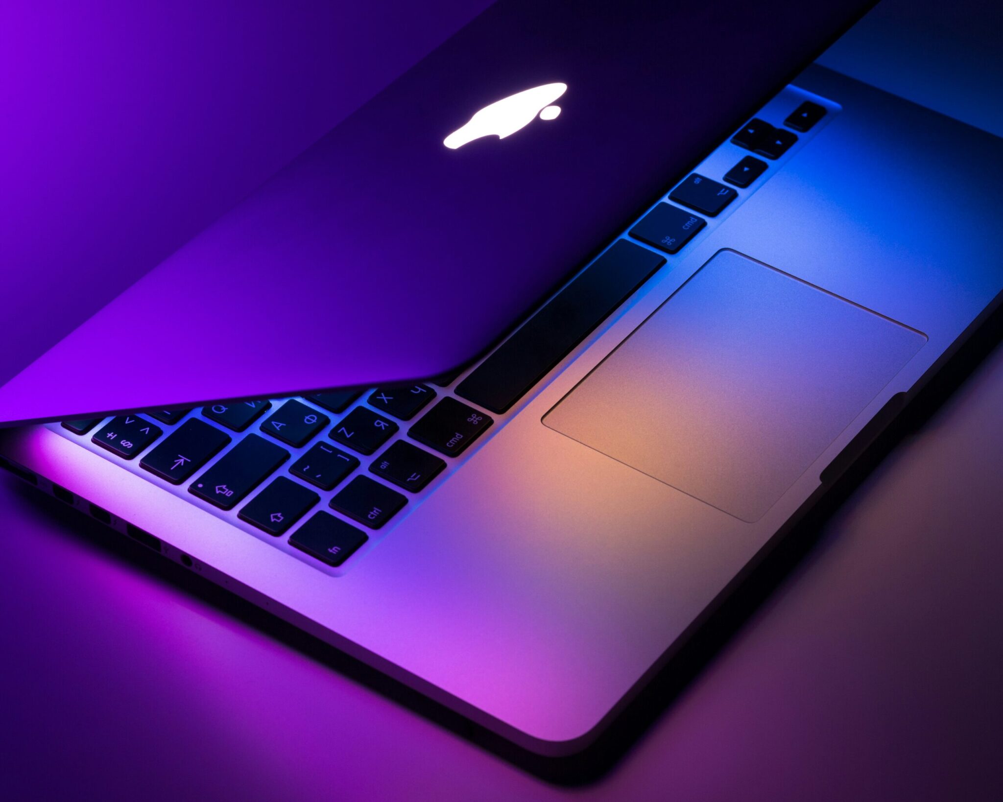 A partially open MacBook laptop with illuminated keyboard and trackpad, featuring purple and orange lighting reflections—showcasing the expert touch of Hardy Repairs.