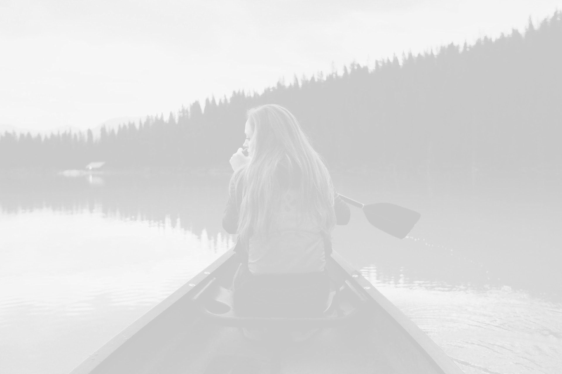 A person with long hair sits in a canoe on a calm lake, surrounded by trees, holding a cup, with a paddle resting beside them.