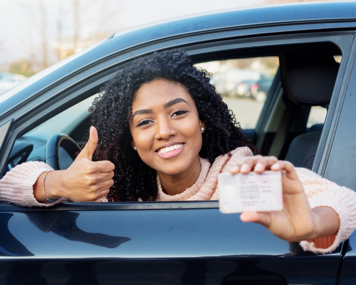 A person sitting in a car smiles, gives a thumbs up, and holds a driver's license out the window after completing driving lessons with Mutlows Driving School.