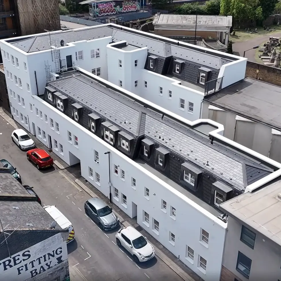 Aerial view of a modern, white apartment building with multiple floors and dormer windows, surrounded by parked cars on a narrow street—a glimpse behind the scenes (BTS) of urban Special Works in progress.