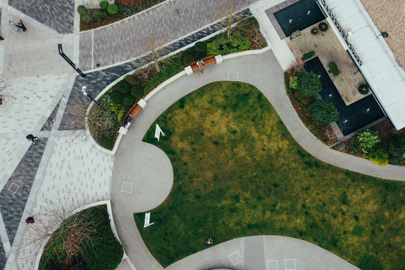 Aerial view of a landscaped urban area featuring curved walkways, green grass, benches, nearby buildings with patterned pavement—an inviting space often considered for Avis Contracts site enhancements.