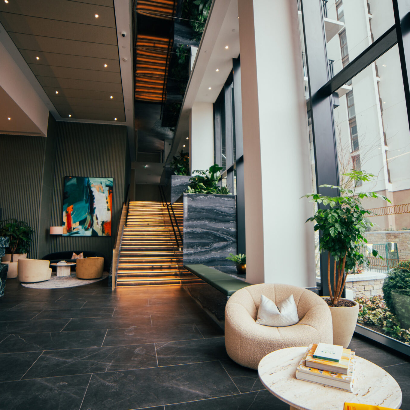 Modern hotel lobby with marble floors, large windows, beige chairs, potted plants, a lit staircase, and contemporary artwork on the wall—ideal for business travelers finalizing Avis Contracts in style.