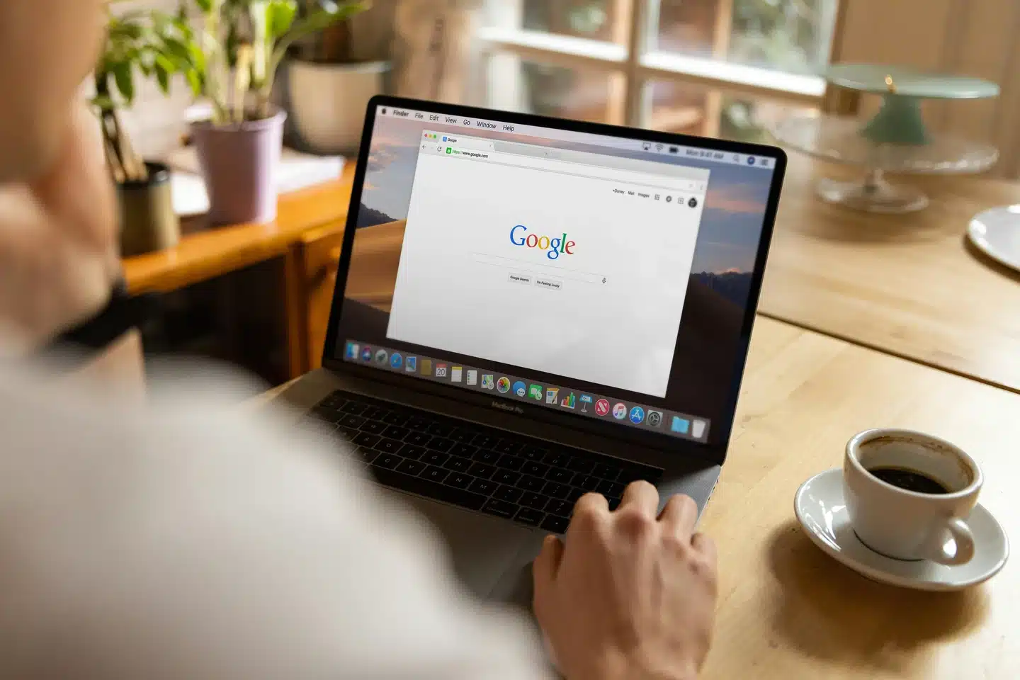 Person using a laptop on a wooden table, displaying the Google homepage. A cup of coffee, plants, and a plate are visible in the background—perfect setting for diving into SEO or planning your next web design project.