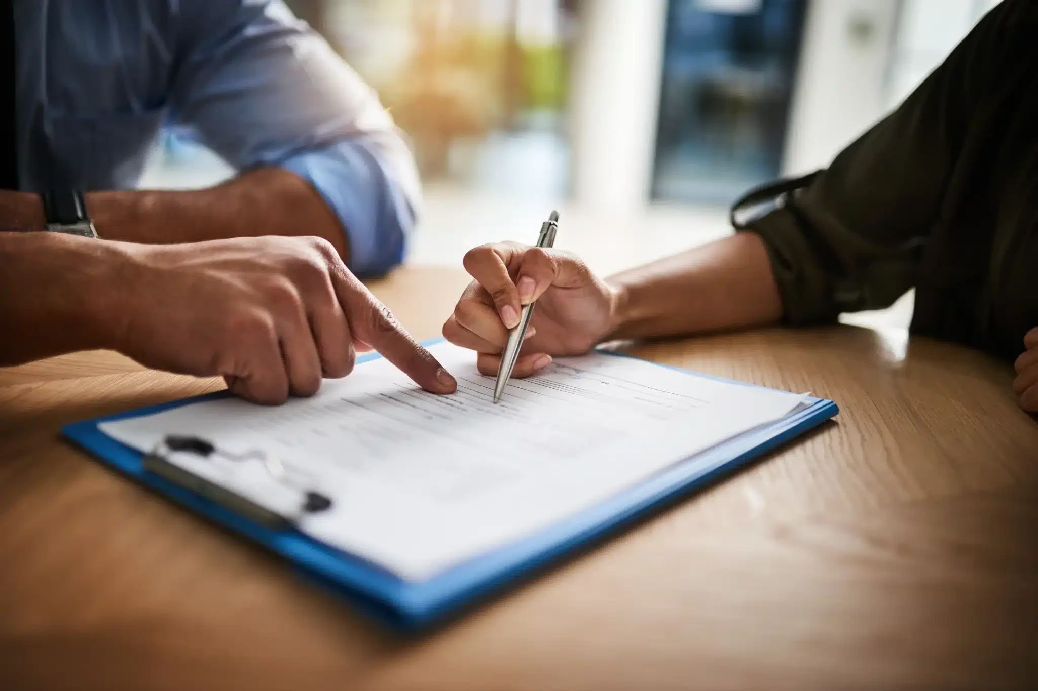 Two people sit at a table, one person points at a document on a clipboard whilst the other holds a pen and prepares to sign, guided by their Compliance Assistant for seamless compliance management.