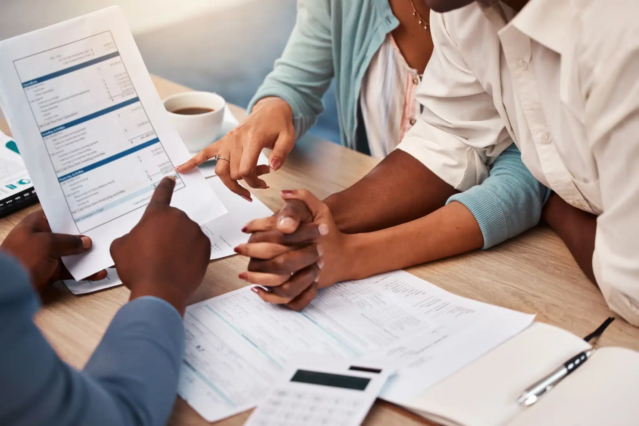Three people sit at a table reviewing financial documents, with a Compliance Assistant visible among the hands, papers, and calculator in close proximity.